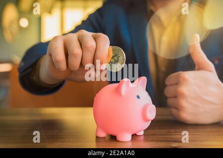 La mano di un uomo d'affari tiene una criptovaluta bitcoin dorata vicino ad una banca piggy del maiale. Bitcoin, criptovaluta e concetto di risparmio. Foto Stock