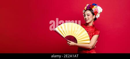 Bella donna asiatica che indossa tradizionale cheongsam abito qipao con colorato fiori sulla testa che tiene il ventilatore dorato in studio isolato rosso banner backgro Foto Stock