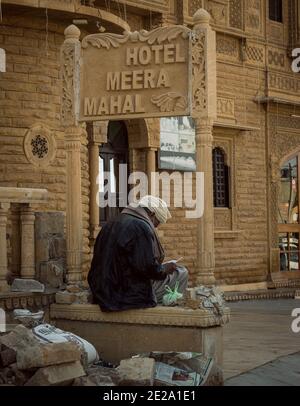 Jaisalmer, Rajasthan / India - 8 gennaio 2021 : vecchio uomo di stile che indossa il turbante e la maschera impropria del viso siede sotto un tabellone scolpito da pietra Foto Stock