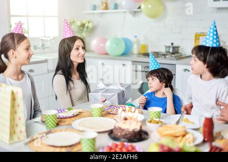 Allegra madre latina di mezza età che tiene una scatola regalo, parlando con i suoi bei bambini mentre si cena, festeggiando il compleanno insieme a casa. Genitorialità, concetto di celebrazione. Messa a fuoco selettiva Foto Stock