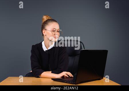 Una donna d'affari carina con gli occhiali siede nell'ufficio e tipi su un calcolatore funzionante. Sfondo grigio. Foto Stock