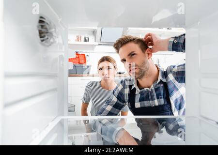 premuroso handyman e giovane donna che guarda il freezer su offuscato primo piano in cucina Foto Stock