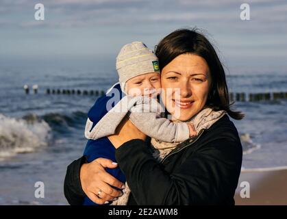 SARBINOWO, POLONIA - 15 ottobre 2017: Donna caucasica polacca felice che tiene un bambino piccolo ragazzo in spiaggia in una giornata nuvolosa Foto Stock