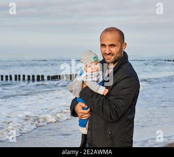 SARBINOWO, POLONIA - 15 ottobre 2017: Uomo che tiene un bambino vicino all'acqua in una spiaggia in una giornata nuvolosa Foto Stock