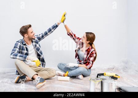 tutta la lunghezza della giovane coppia felice in guanti che danno ciascuno altri cinque alti mentre si siedono sul pavimento vicino ai barattoli di dipingi a casa Foto Stock