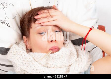 una giovane bella ragazza è malata a casa a letto, tiene la sua fronte calda e misura la temperatura con un termometro Foto Stock