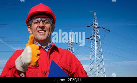 Sorridente lavoratore di servizi pubblici elettrici che dà Thumbs in su vicino alle torri di trasmissione elettriche. Ritratto di sorridente ingegnere in Red Hardhat e tute. Foto Stock