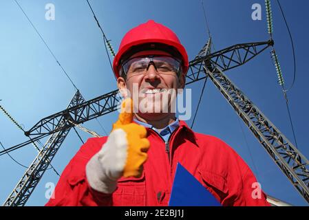 Operatore di servizi elettrici che fornisce le miniature in alto accanto alla torre di trasmissione elettrica. Ritratto di sorridente ingegnere in Red Hardhat e tute. Foto Stock