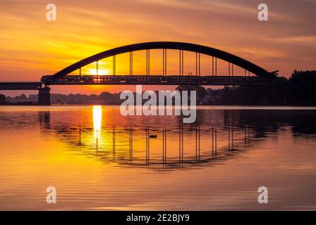 Bilevel / treno interurbano a due piani dalla Nederlandse Spoorwegen che cavalcano il ponte ferroviario ad arco, squaleggiato contro l'alba, nei Paesi Bassi Foto Stock