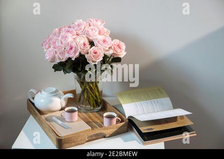 Rosa Bianco Rosa o'hara. Bouquet di rose rosa in vaso di vetro con una teiera bianca e due tazze di cappuccino su un vassoio su un tavolino tra i libri. Foto Stock