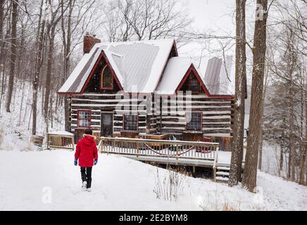 Ragazzo in cappotto rosso invernale che cammina verso la cabina di tronchi in giornata invernale innevata. Foto Stock