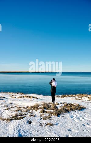 vista posteriore di una donna irriconoscibile viaggiatore in abiti caldi e. cappello in piedi sulla costa innevata di mare calmo e ammirando natura contro cielo senza nuvole Foto Stock