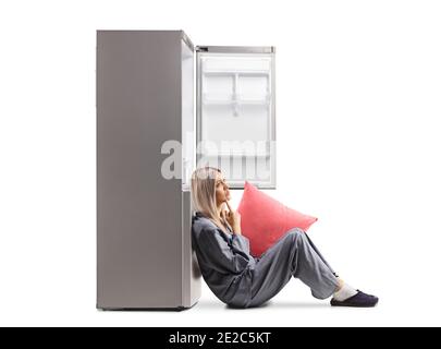 Giovane donna in pigiama seduta sul pavimento e appoggiata un frigorifero vuoto isolato su sfondo bianco Foto Stock