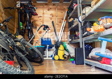 Unità di deposito di utilità di legno della casa suburbana con roba vario su mensole, biciclette, macchina di esercitazione, scala, attrezzi di giardino e attrezzatura. Disordinato Foto Stock