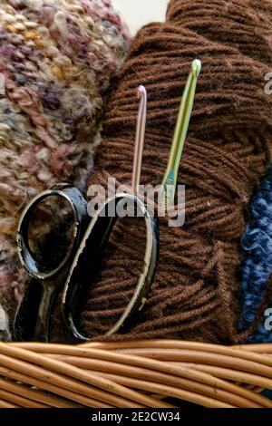 Ganci a crochet in alluminio verde e rosa e forbici nere indossate mostrate in un cestello di vimini con matasse di filato marrone e multicolore. Foto Stock