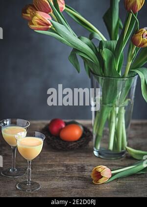 Uova di Pasqua colorate in nido di vimini, bicchiere di uovo di nog e tulipani in vaso su tavola di legno. Spazio di copia. Concetto di vacanza di Pasqua. Foto Stock