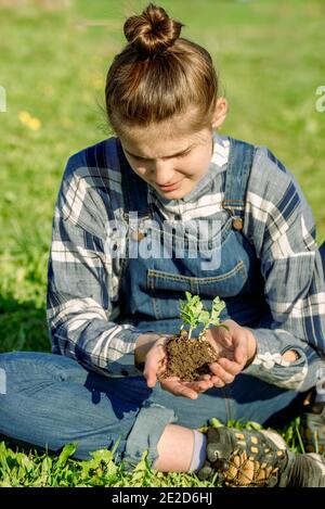 Coltivatore di ragazza in tute di denim tiene un germoglio con terra. Piantando verdure. Concetto di conservazione dell'ambiente Foto Stock