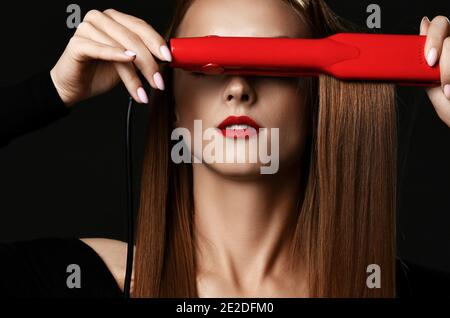 Giovane bella donna con lunghi capelli lisci setosi in nero vestiti che coprono gli occhi con piastra per capelli rossa Foto Stock