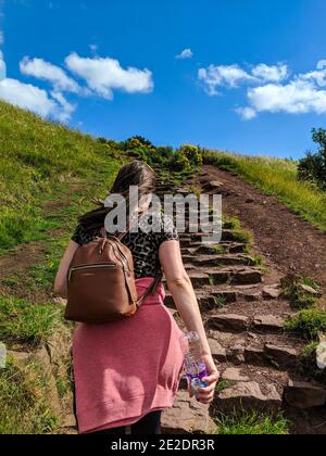 Una giovane donna di brunette cammina lungo i gradini verso Arthur's Seat, Edimburgo, in una giornata soleggiata e azzurra. Foto Stock