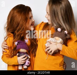 Sorridi piccole ragazze sorelle o amici in maniche lunghe gialle in piedi abbracciando tenendo le bambole giocattolo in mano e guardando ciascuno altro Foto Stock