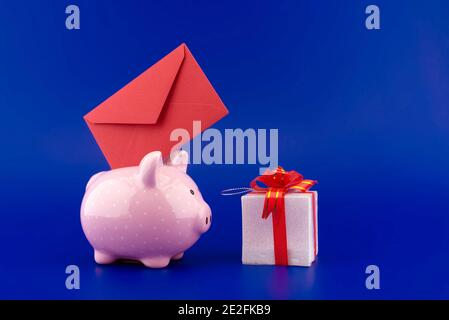 Busta rossa festiva inserita in una banca rosa piggy accanto a una casella presente su sfondo blu con copia spazio Foto Stock