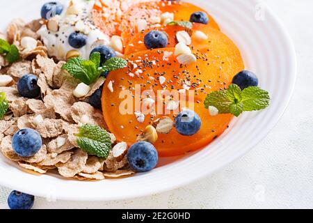 Ciotola di fiocchi con yogurt, blueberryes e persimmon su tavola di legno bianco. Cibo fitness. Foto Stock
