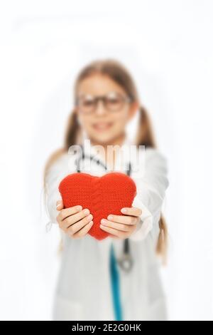 Cute ragazza vestita come medico tiene un cuore giocattolo rosso davanti a lei. Concetto di salute del cuore del bambino Foto Stock