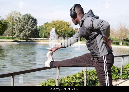 Uomo nero africano con cuffie che allunga la gamba in un parco. Fitness e sport all'aperto. Foto Stock