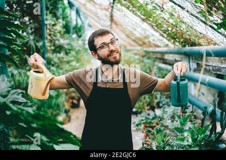 Giovane uomo felice giardiniere ambientalista che tiene in mano una lattina di irrigazione, curando per le piante in serra. Foto Stock