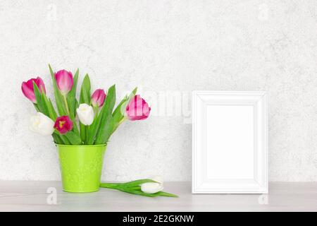 Cornice bianca vuota con bouquet di fiori di tulipano sul tavolo vicino alla parete. Messa a fuoco selettiva. Sfondo mock-up primaverile Foto Stock