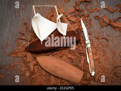 Barca giocattolo fatta di corteccia di quercia. Piccola nave a vela in legno. Sullo sfondo un coltello da tasca e trucioli, lavoro in corso. Foto Stock