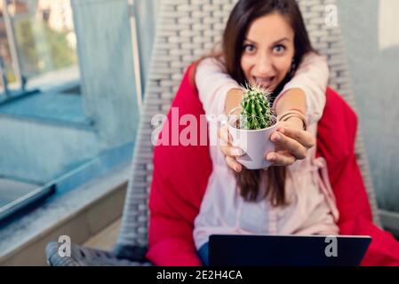 Giovane donna seduta su un balcone su un altalena e. tenere pentola con cactus che punta alla macchina fotografica e fare Face Funny Focus sul cactus Foto Stock