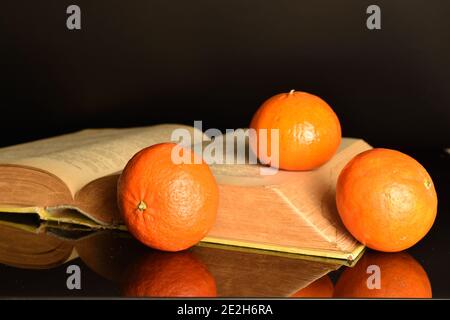 Tre interi tondi arancio brillante succosa mandarino maturo giace su un libro aperto, su uno sfondo nero. Foto Stock