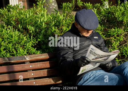 Uomo in berretto piatto seduto su banco di lettura giornale Foto Stock