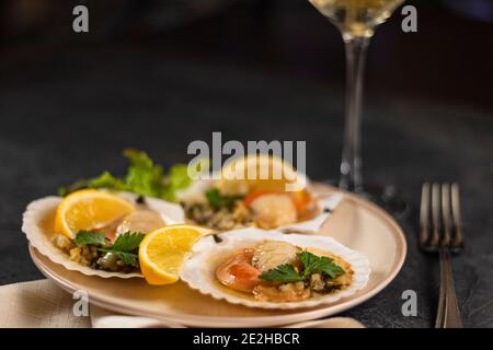 Capesante con limone su fondo nero con tovagliolo e forchetta. Capesante cotte al forno con caviale in un piatto sullo sfondo di bicchieri sfocati di Foto Stock