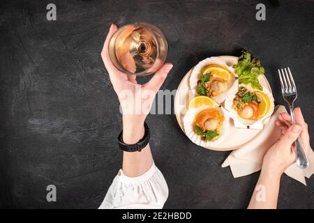 Capesante al forno con caviale e un bicchiere di vino su fondo nero. Le mani femminili tengono un bicchiere di vino bianco e una forchetta accanto alle capesante cotte con c Foto Stock