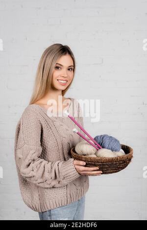 Giovane donna che sorride alla macchina fotografica mentre tiene il cesto con la maglia aghi e filati su sfondo bianco Foto Stock