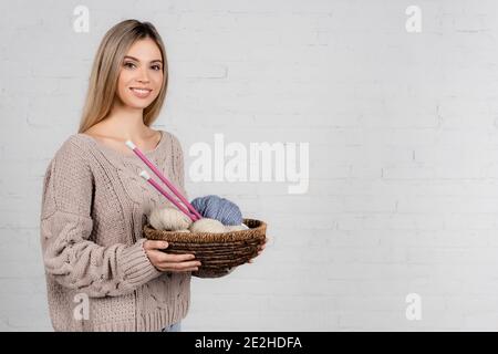 Donna sorridente che tiene il cesto con aghi da maglieria e filo di lana palline su sfondo bianco Foto Stock