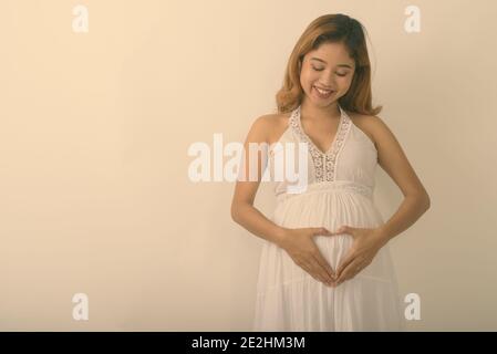 Studio shot di giovani felici Asian incinta Donna sorridente mentre si effettua a mano segno di cuore sul suo stomaco con amore per il suo bambino contro uno sfondo bianco Foto Stock