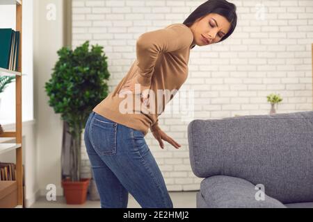 La giovane donna si alza dal divano e si sente improvvisamente intensa dolore nella parte inferiore della schiena Foto Stock