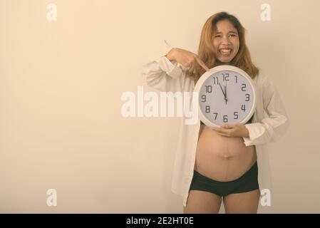 Studio shot di giovani asiatici felice donna incinta sorridere mentre puntando al muro orologio contro uno sfondo bianco Foto Stock