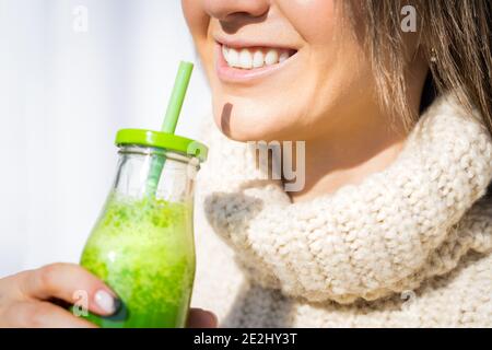 Donna sorridente in pullover di lana che beve frullato disintossicazione o succo di frutta fatti in casa verde. Mangiare sano e pulito Foto Stock