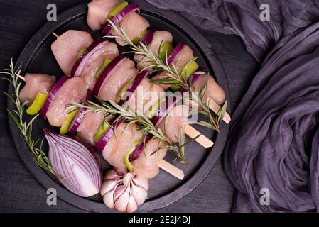 Pollo fresco kebab. Spiedini di pollo crudo su un piatto nero con spezie e rosmarino verde e cipolle su uno sfondo di legno. Pollo crudo . Foto Stock