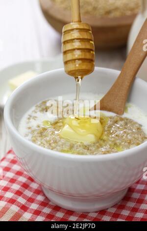 Far cadere il miele con un cucchiaino di legno sul porridge scozzese con latte e burro. Foto Stock