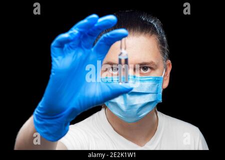 il medico in maschera protettiva e guanti sterili contiene una fiala di vetro con il vaccino contro il virus in mano e guarda valuta attentamente il prod medico Foto Stock