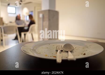 Piatto bianco vuoto e sporco con cucchiaio e forchetta in acciaio inossidabile, e sul tavolo nero dopo il pasto finale, e sfondo di cucina sfocata, mensa dell'ufficio. Foto Stock