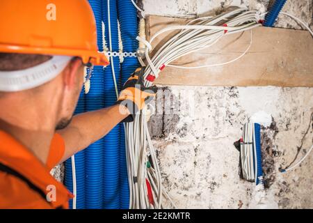 Orange Hard Hat indossando un elettricista caucasico nel suo mazzo di cavi elettrici in movimento anni '40 all'interno di un edificio commerciale di recente sviluppo. Costruzione e. Foto Stock