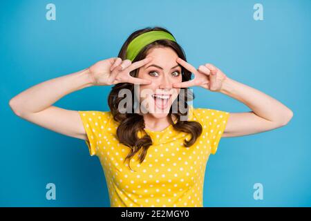 Ritratto di attraente ragazza eccitata fare v-segno vicino occhi indossare polka-dot vestiti isolati su sfondo blu Foto Stock