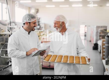 Immagine di due dipendenti in abiti sterili in fabbrica di alimenti sorridenti e parlanti. L'uomo maturo sta tenendo il vassoio pieno di biscotti freschi mentre il più giovane è Foto Stock
