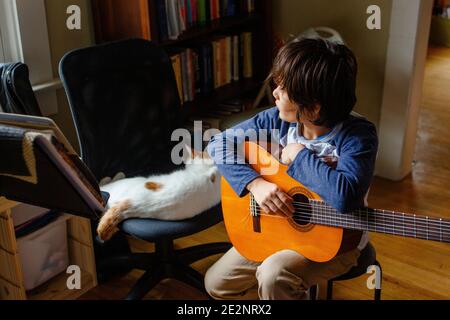 Un ragazzo guarda fuori una finestra che tiene una chitarra accanto ad un gatto che dorme Foto Stock